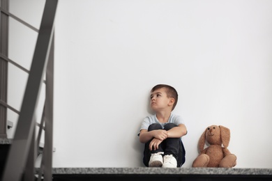 Sad little boy with toy sitting near white wall