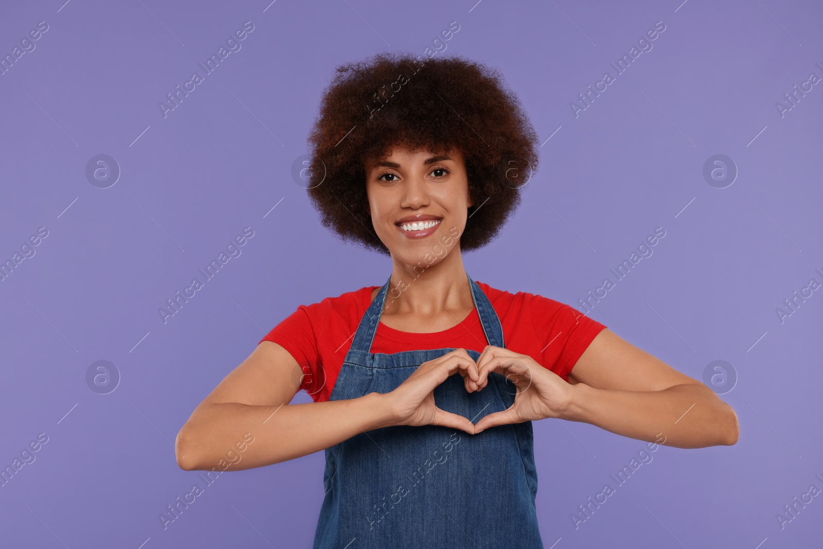 Photo of Happy young woman in apron showing heart gesture on purple background