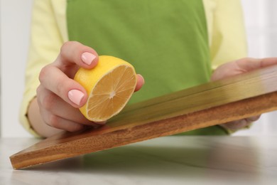 Photo of Woman rubbing wooden cutting board with lemon at white table, closeup