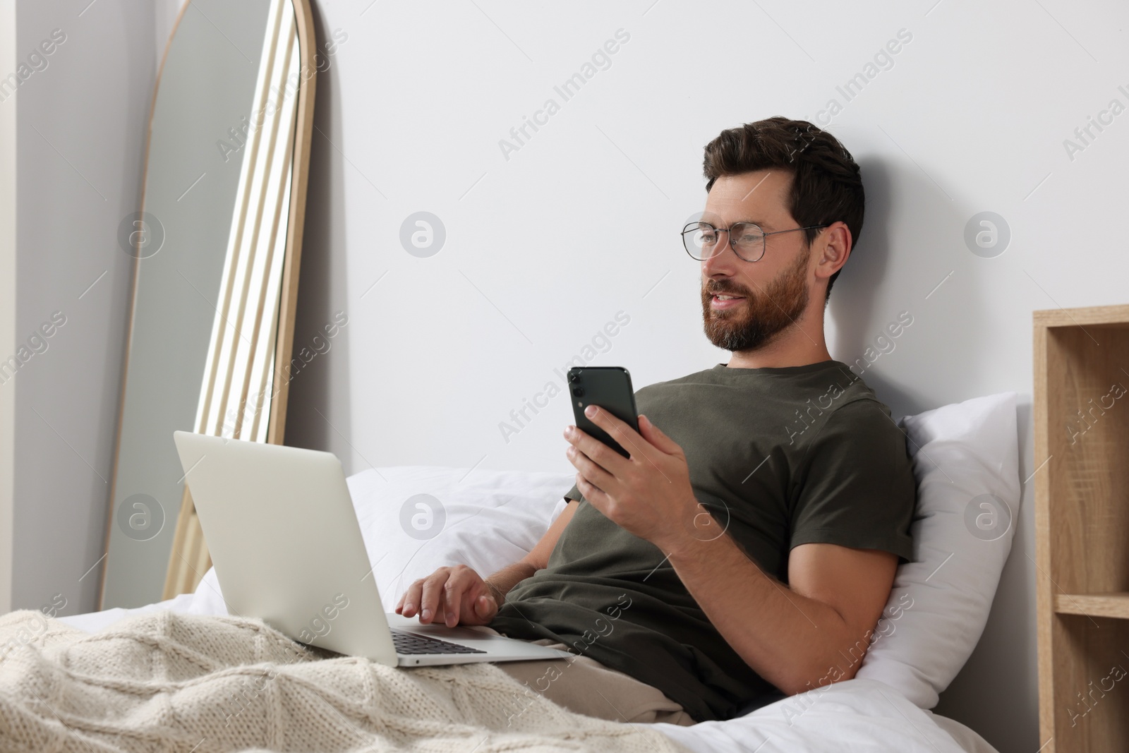 Photo of Handsome man using smartphone and laptop in bed at home