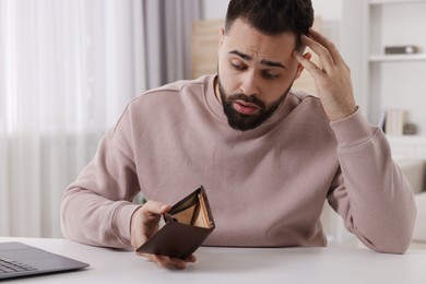 Confused man with empty wallet at white table indoors