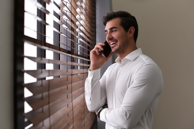 Photo of Handsome young man talking on smartphone near window indoors