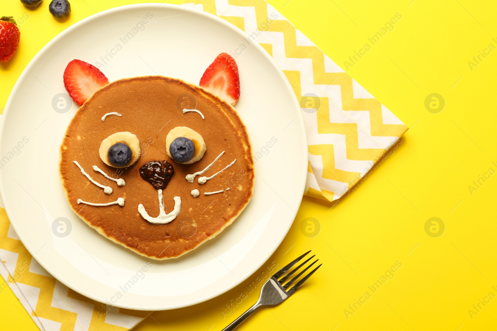 Photo of Creative serving for kids. Plate with cute cat made of pancakes, berries, cream, banana and chocolate paste on yellow background, flat lay