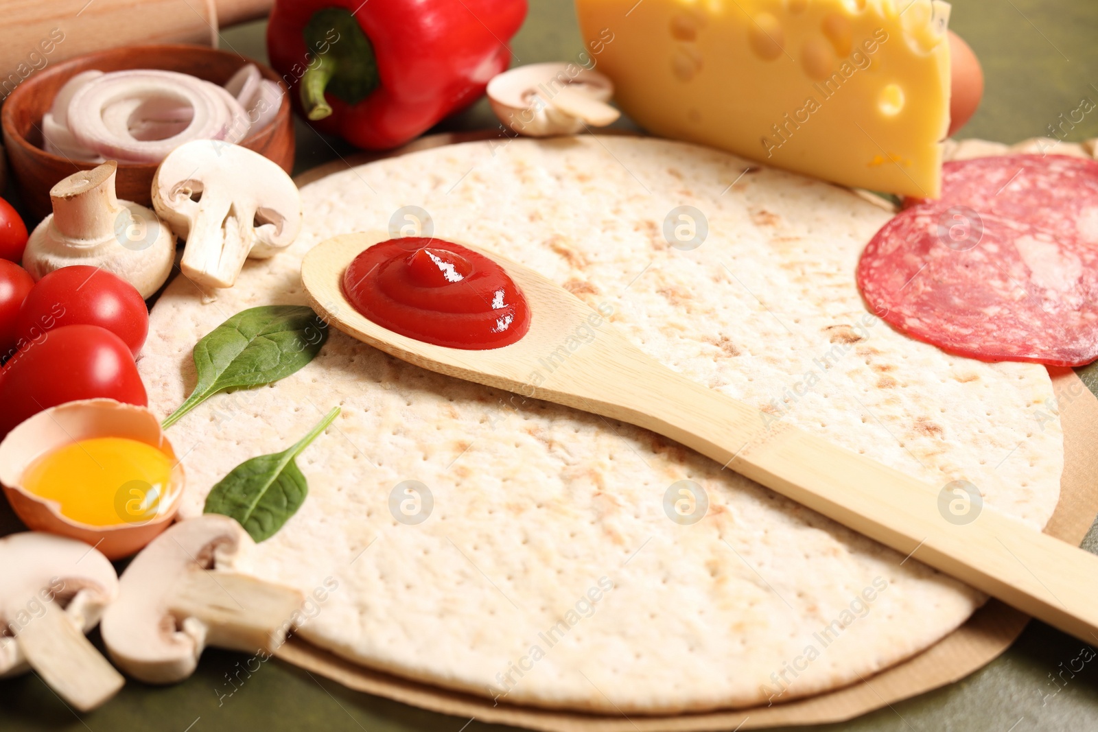 Photo of Pizza base, spoon with tomato sauce and products on green table, closeup