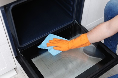 Photo of Young man cleaning oven with rag in kitchen, closeup