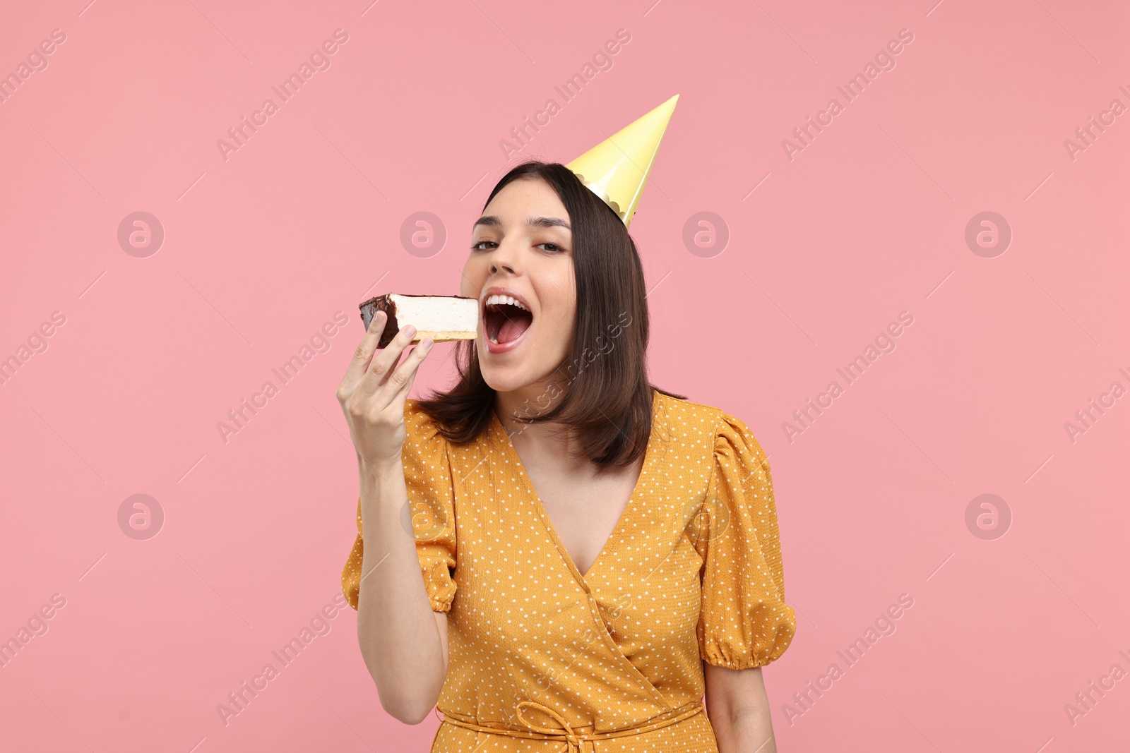 Photo of Young woman in party hat eating cheesecake on pink background