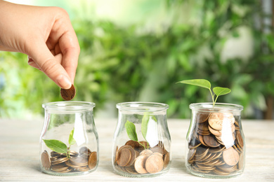 Woman putting coin into jar with green plant on white wooden table, closeup