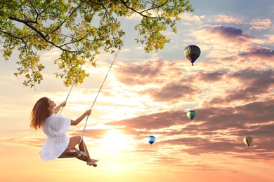Image of Dream world. Young woman swinging, hot air balloons in sunset sky on background