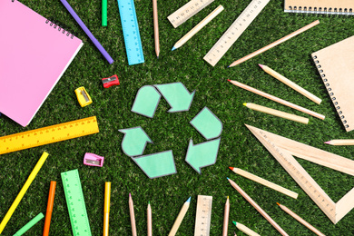 Recycling symbol, plastic and wooden stationery on green grass, flat lay