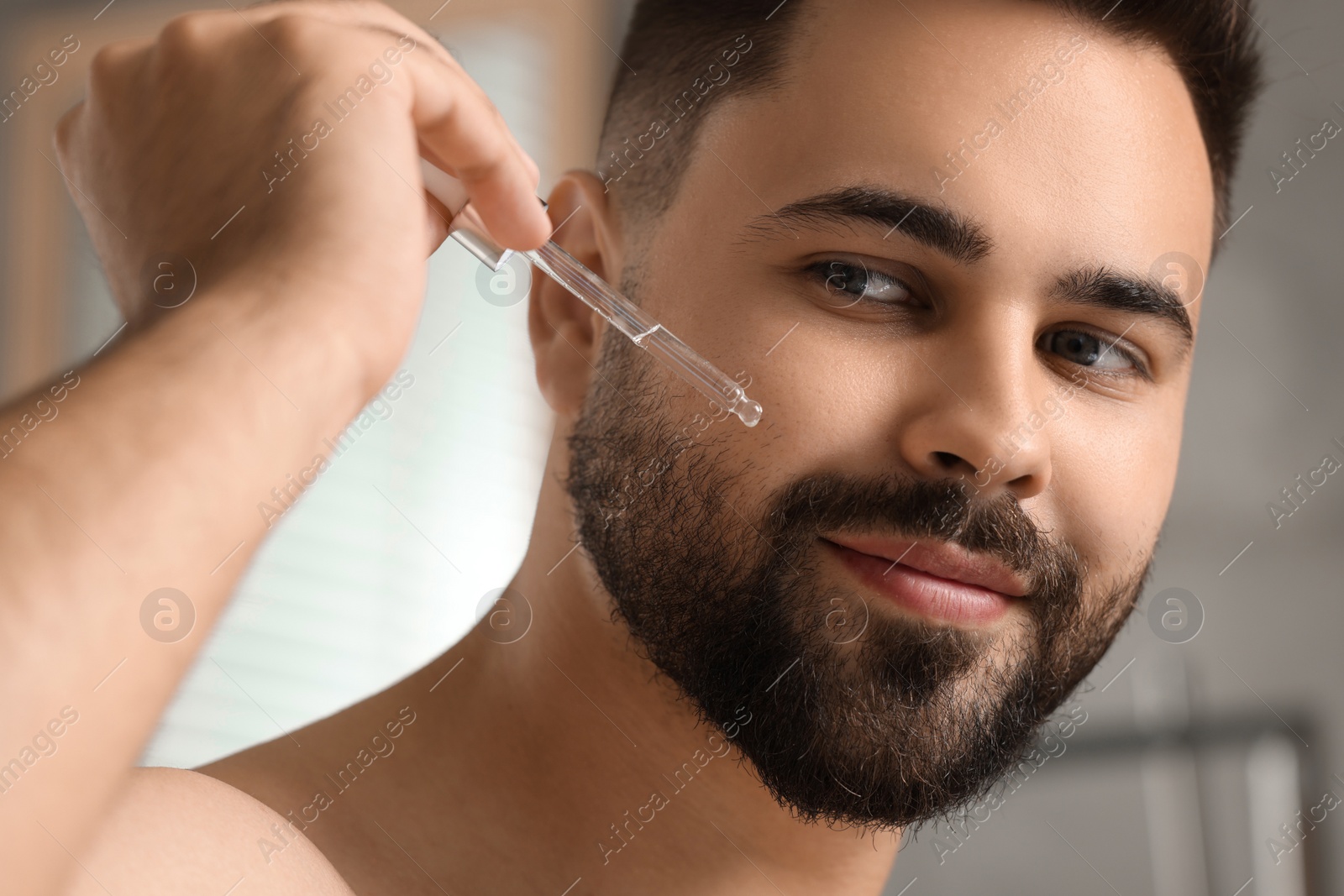 Photo of Handsome man applying cosmetic serum onto his face on blurred background, closeup