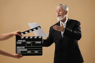 Photo of Senior actor performing role while second assistant camera holding clapperboard on beige background, selective focus. Film industry