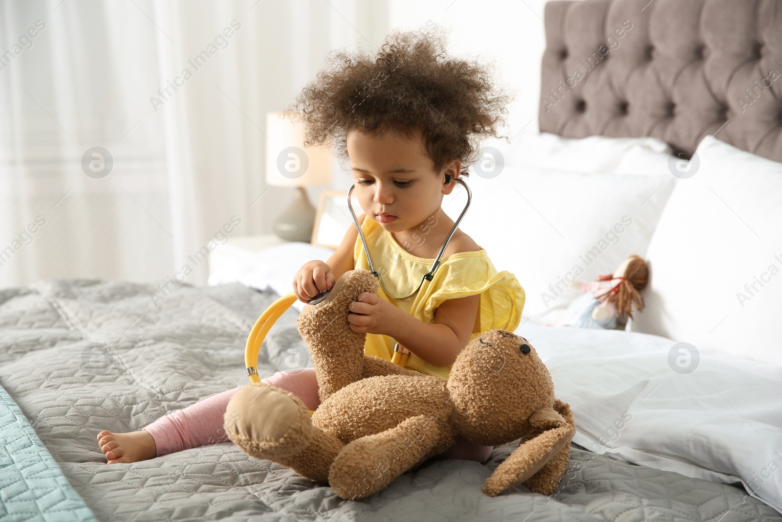 Photo of Cute African American child imagining herself as doctor while playing with stethoscope and toy bunny at home