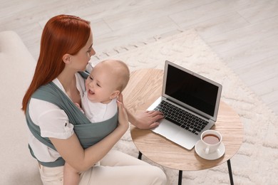Mother using laptop while holding her child in sling (baby carrier) indoors