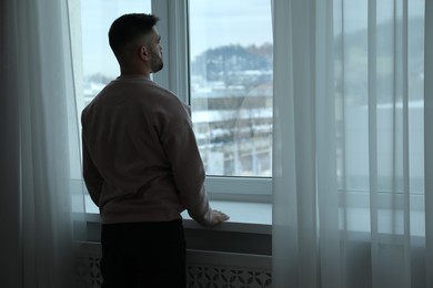 Photo of Sad man looking at window at home, back view. Space for text