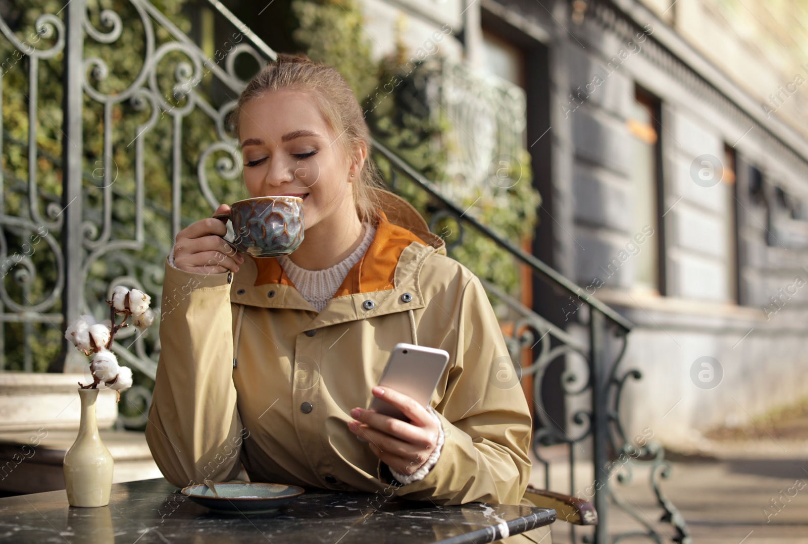 Photo of Young woman enjoying tasty coffee while using mobile phone at table outdoors