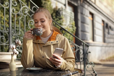 Photo of Young woman enjoying tasty coffee while using mobile phone at table outdoors