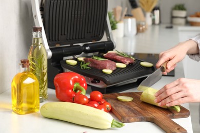 Photo of Woman cooking different products with electric grill at white wooden table in kitchen, closeup