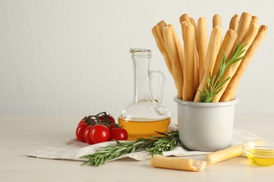 Photo of Delicious grissini sticks, oil, rosemary and tomatoes on white wooden table. Space for text