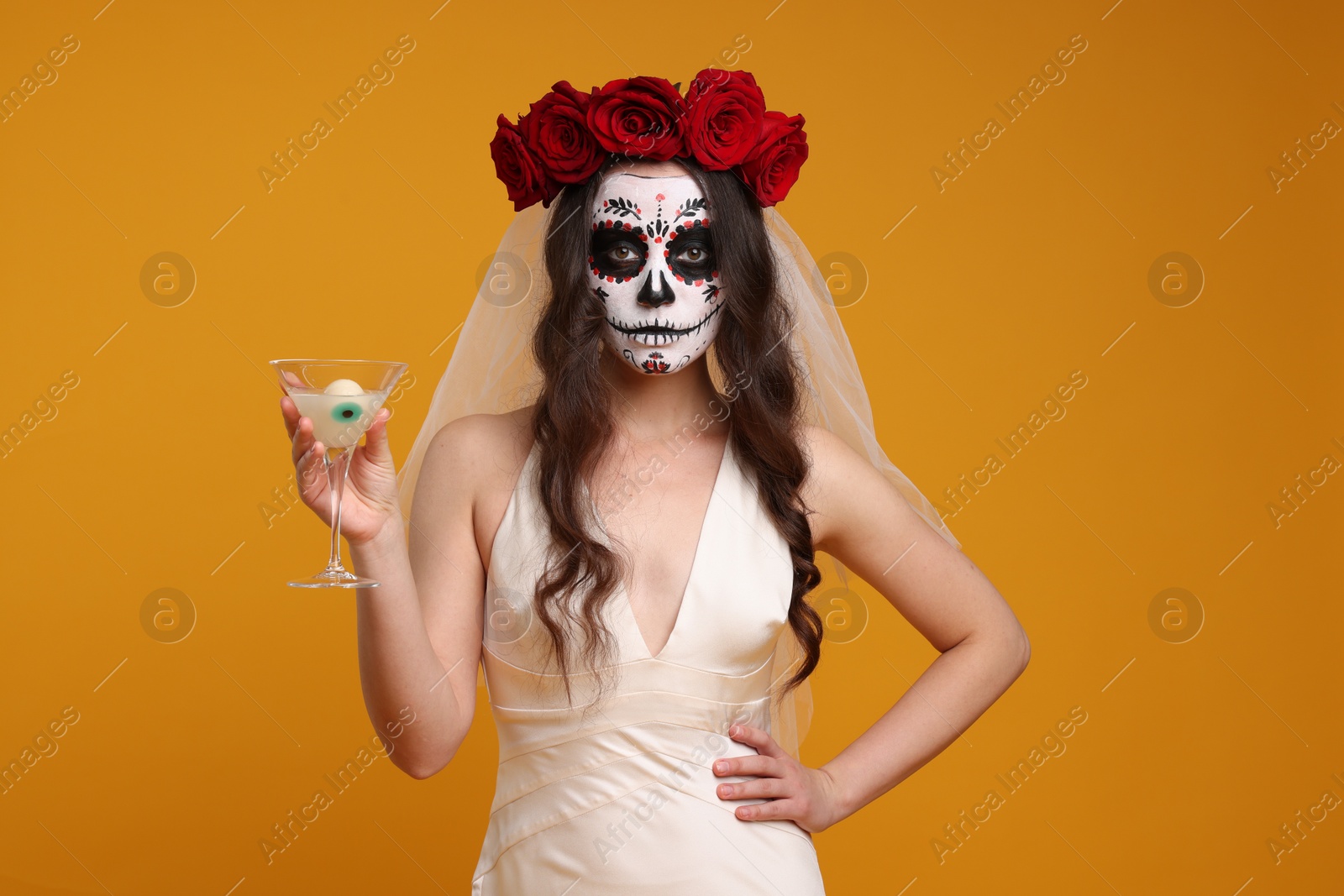 Photo of Young woman in scary bride costume with sugar skull makeup, flower crown and cocktail on orange background. Halloween celebration