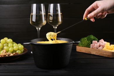 Photo of Woman dipping grape into fondue pot with melted cheese at black wooden table, closeup