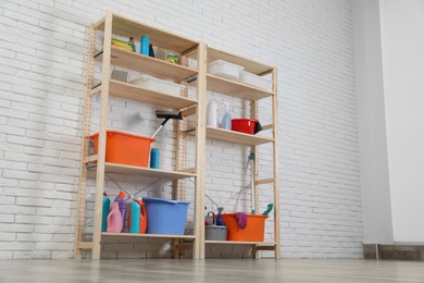 Wooden shelving units with cleaning equipment near white brick wall. Stylish room interior