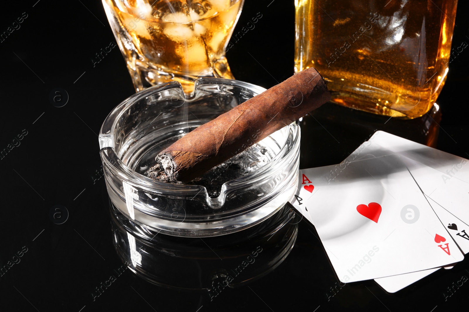 Photo of Smoldering cigar, ashtray, playing cards and whiskey on black mirror surface, closeup