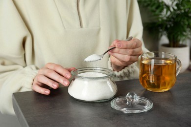 Woman adding sugar into aromatic tea at grey table indoors, closeup