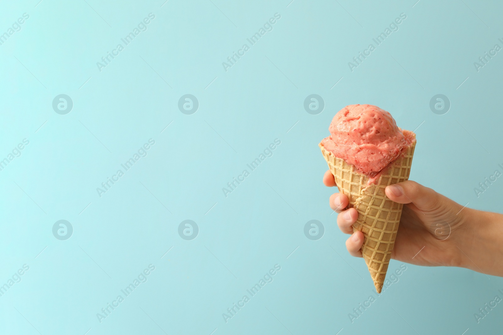 Photo of Woman holding waffle cone with delicious pink ice cream on light blue background, closeup. Space for text