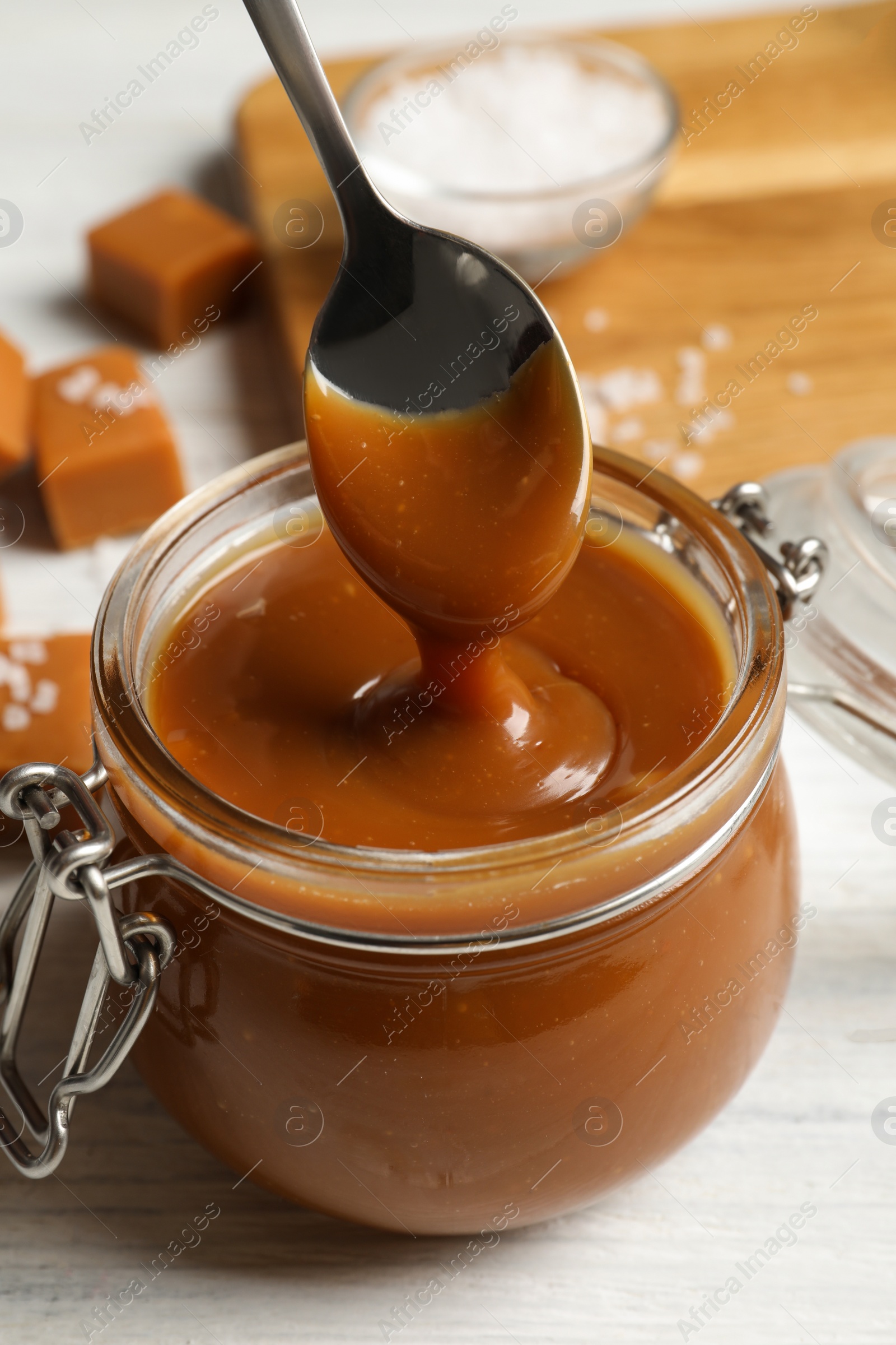 Photo of Taking yummy salted caramel with spoon from glass jar at white wooden table, closeup