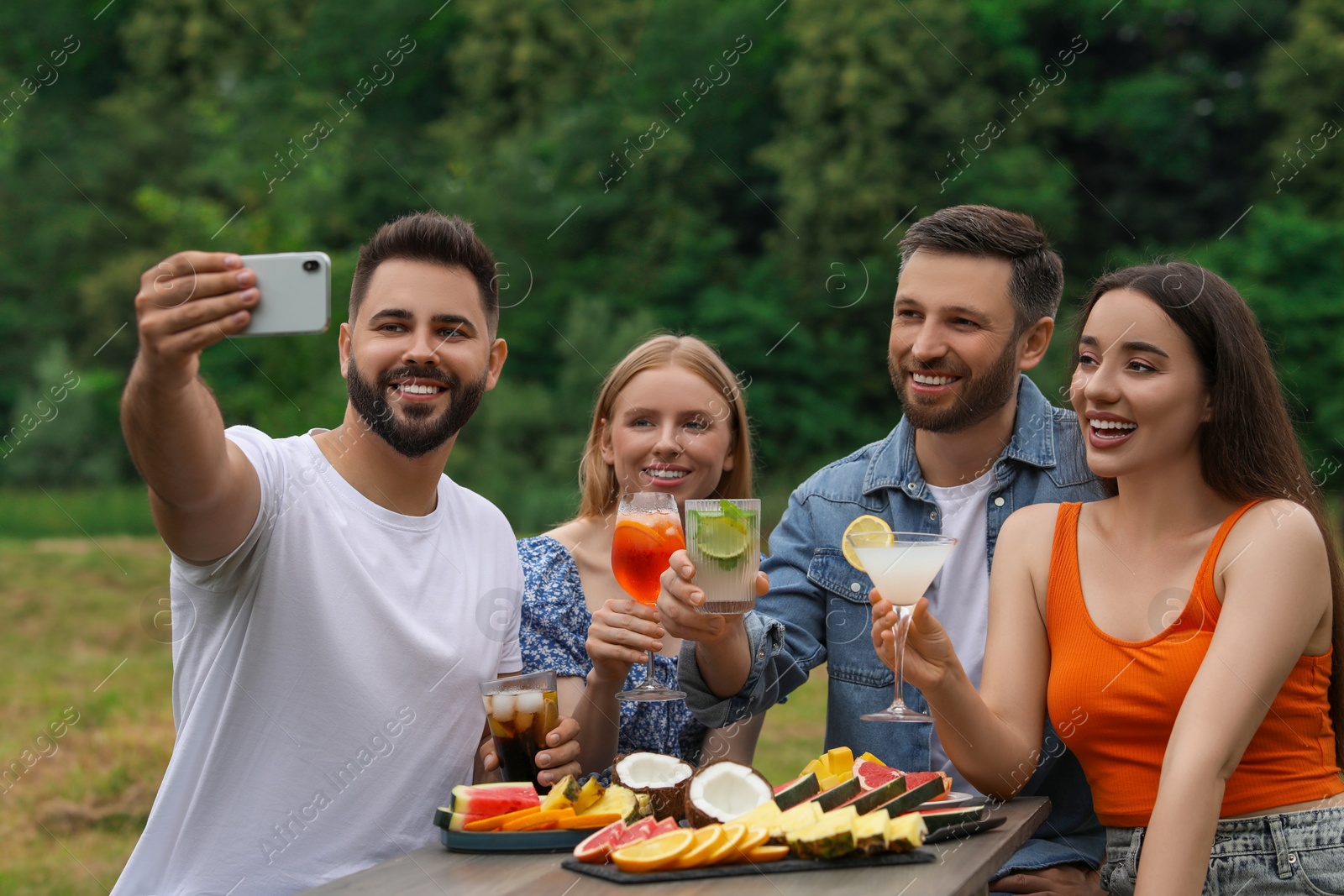 Photo of Happy friends with glasses of cocktails taking selfie at table outdoors