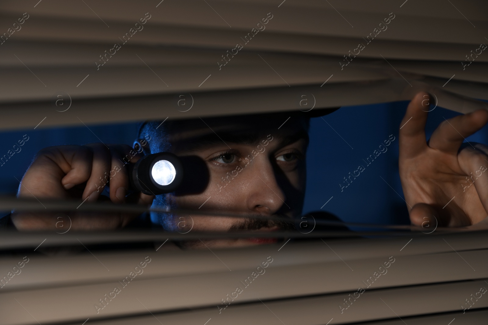 Photo of Male security guard with flashlight looking through window blinds in darkness