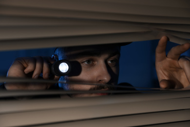 Male security guard with flashlight looking through window blinds in darkness