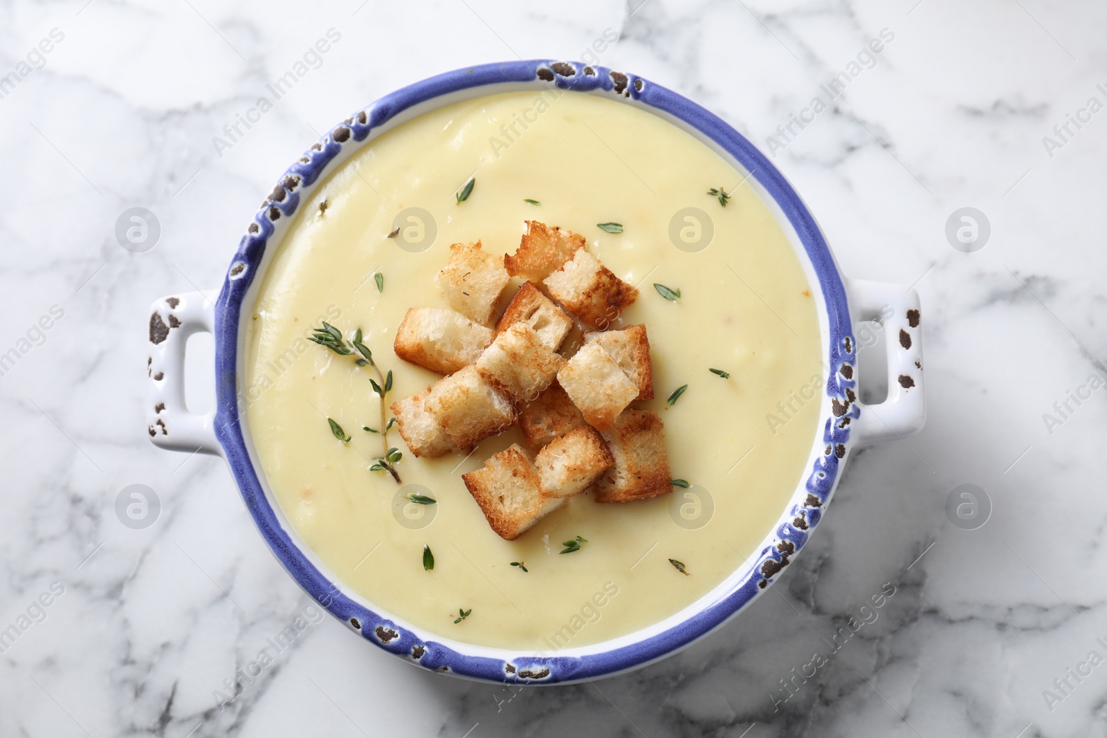 Photo of Tasty potato soup with croutons and rosemary in ceramic pot on white marble table, top view