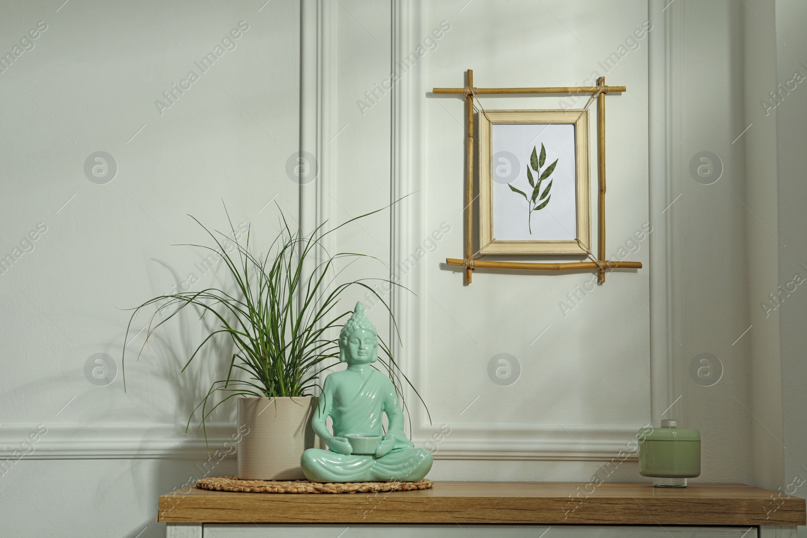Photo of Potted houseplant, buddha statue and candle in jar on wooden table near white wall with stylish bamboo frame