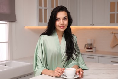 Photo of Pretty young woman wearing beautiful silk robe with cup of coffee in kitchen