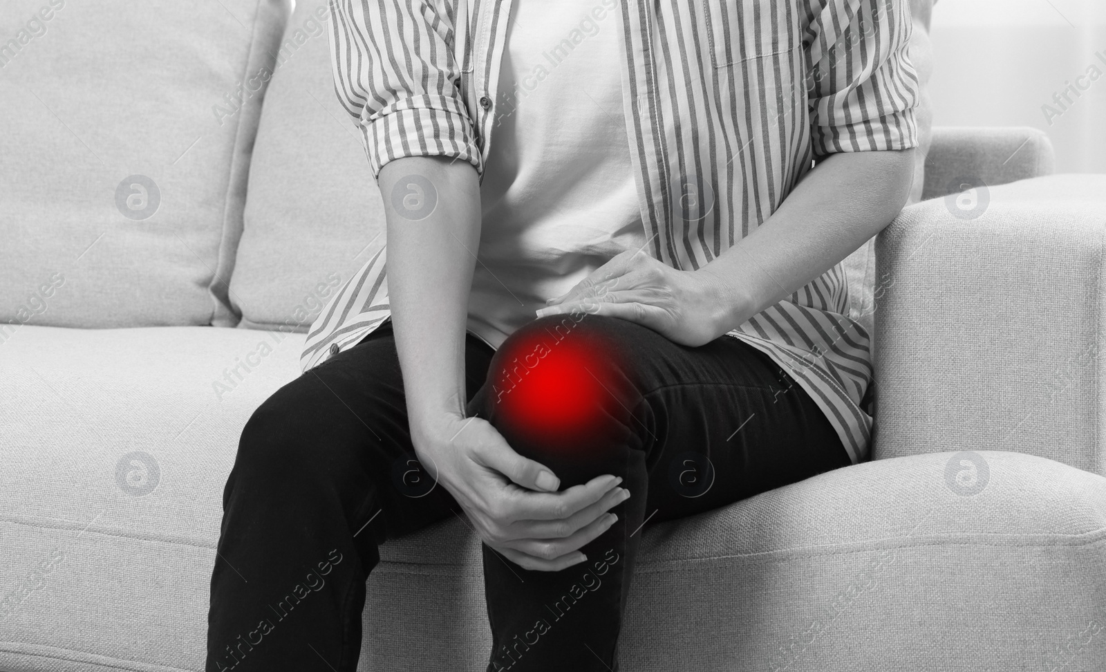 Image of Woman suffering from pain in knee on sofa at home, closeup. Black and white effect