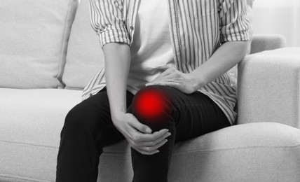 Woman suffering from pain in knee on sofa at home, closeup. Black and white effect