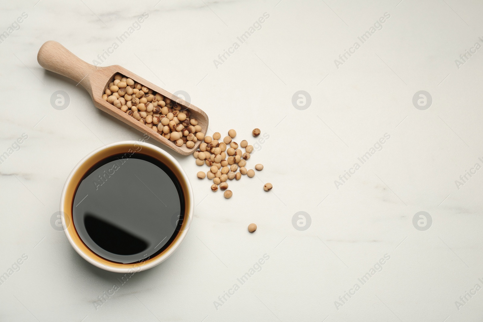 Photo of Soy sauce in bowl and scoop with soybeans on white marble table, flat lay. Space for text