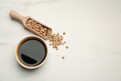 Photo of Soy sauce in bowl and scoop with soybeans on white marble table, flat lay. Space for text