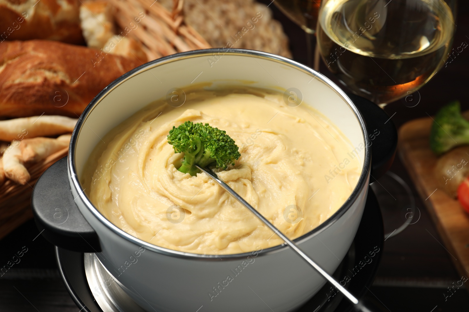 Photo of Dipping piece of broccoli into fondue pot with melted cheese at table, closeup