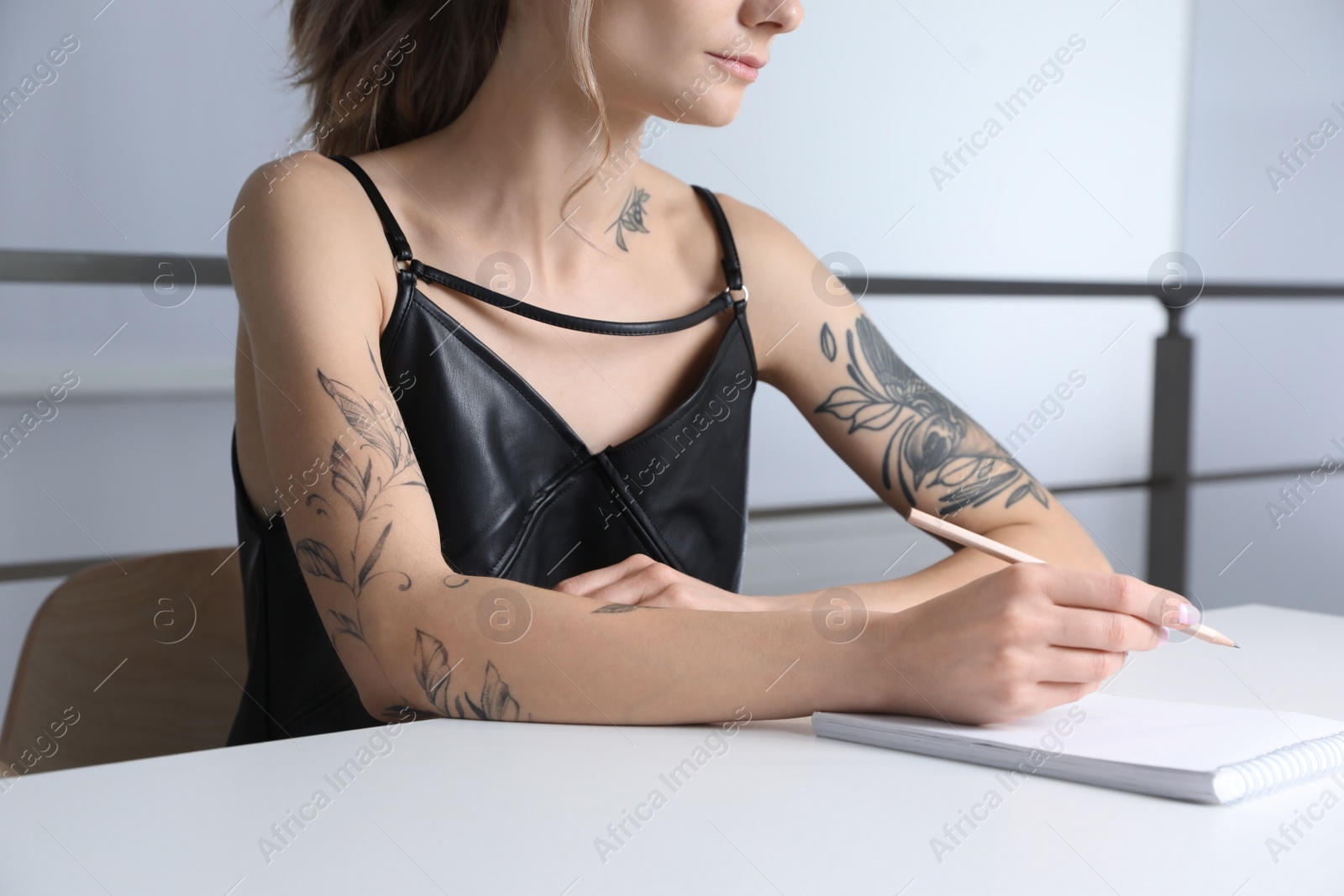 Photo of Beautiful woman with tattoos on body drawing in sketchbook at table indoors, closeup