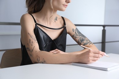 Photo of Beautiful woman with tattoos on body drawing in sketchbook at table indoors, closeup
