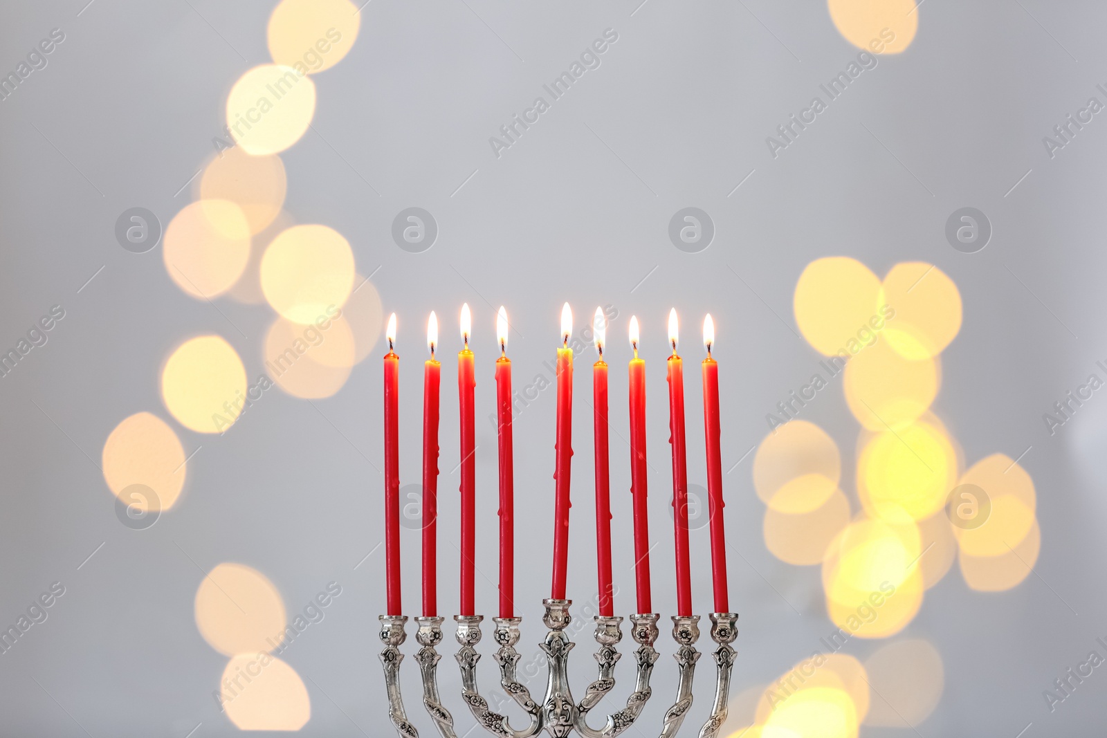 Photo of Silver menorah with burning candles against light grey background and blurred festive lights, space for text. Hanukkah celebration