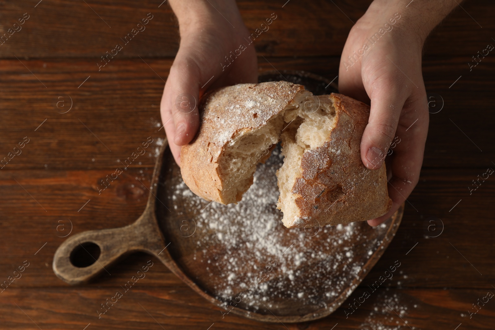 Photo of Man breaking loaf of fresh bread at wooden table, top view