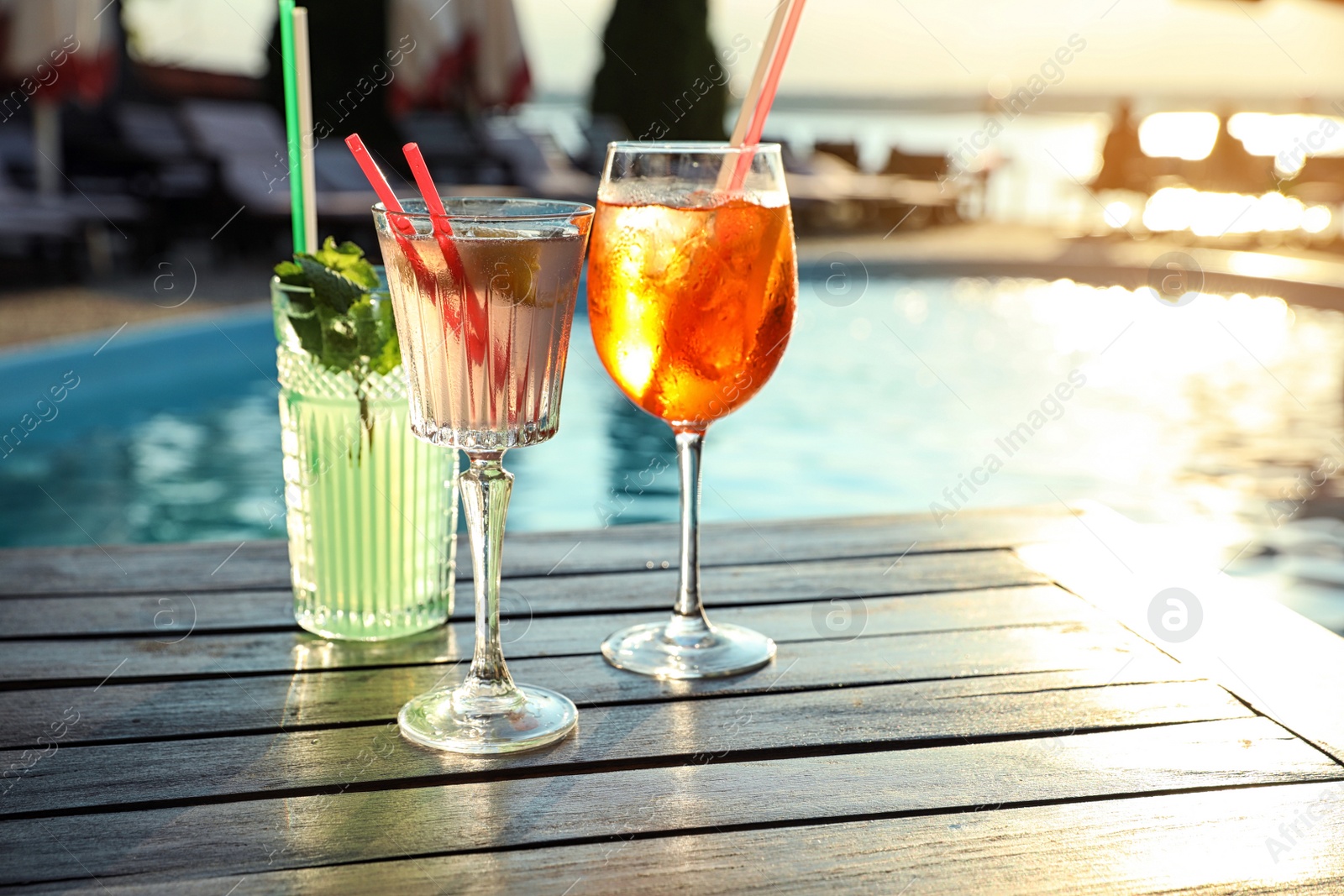Photo of Glasses of fresh summer cocktails on wooden table near swimming pool outdoors at sunset. Space for text