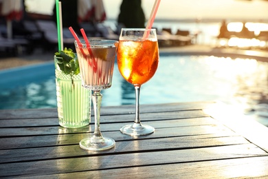 Photo of Glasses of fresh summer cocktails on wooden table near swimming pool outdoors at sunset. Space for text