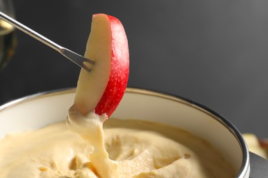 Photo of Dipping piece of apple into fondue pot with melted cheese on grey background, closeup