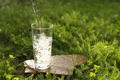 Photo of Pouring fresh water into glass on stone in green grass outdoors. Space for text