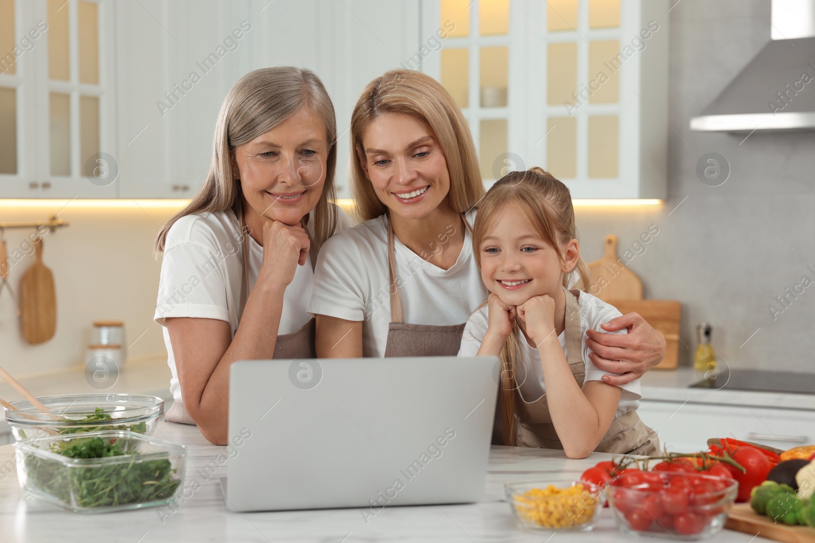 Photo of Three generations. Happy grandmother, her daughter and granddaughter using laptop in kitchen