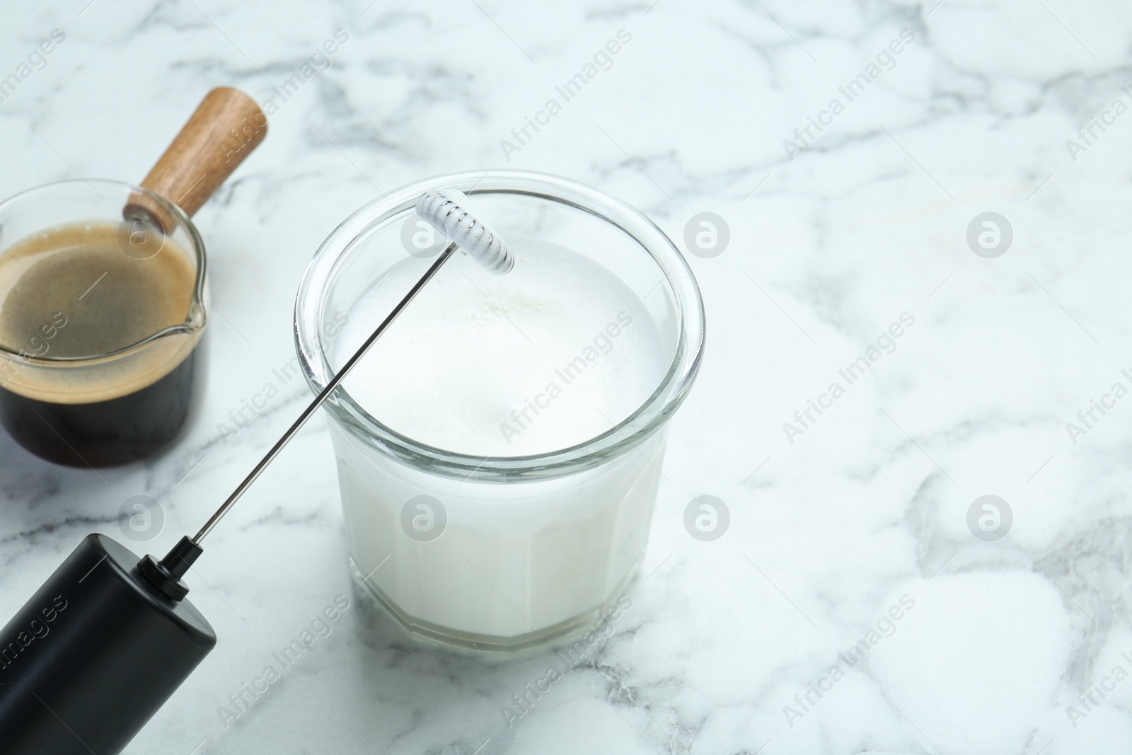 Photo of Mini mixer (milk frother), whipped milk in glass and coffee on white marble table, closeup. Space for text